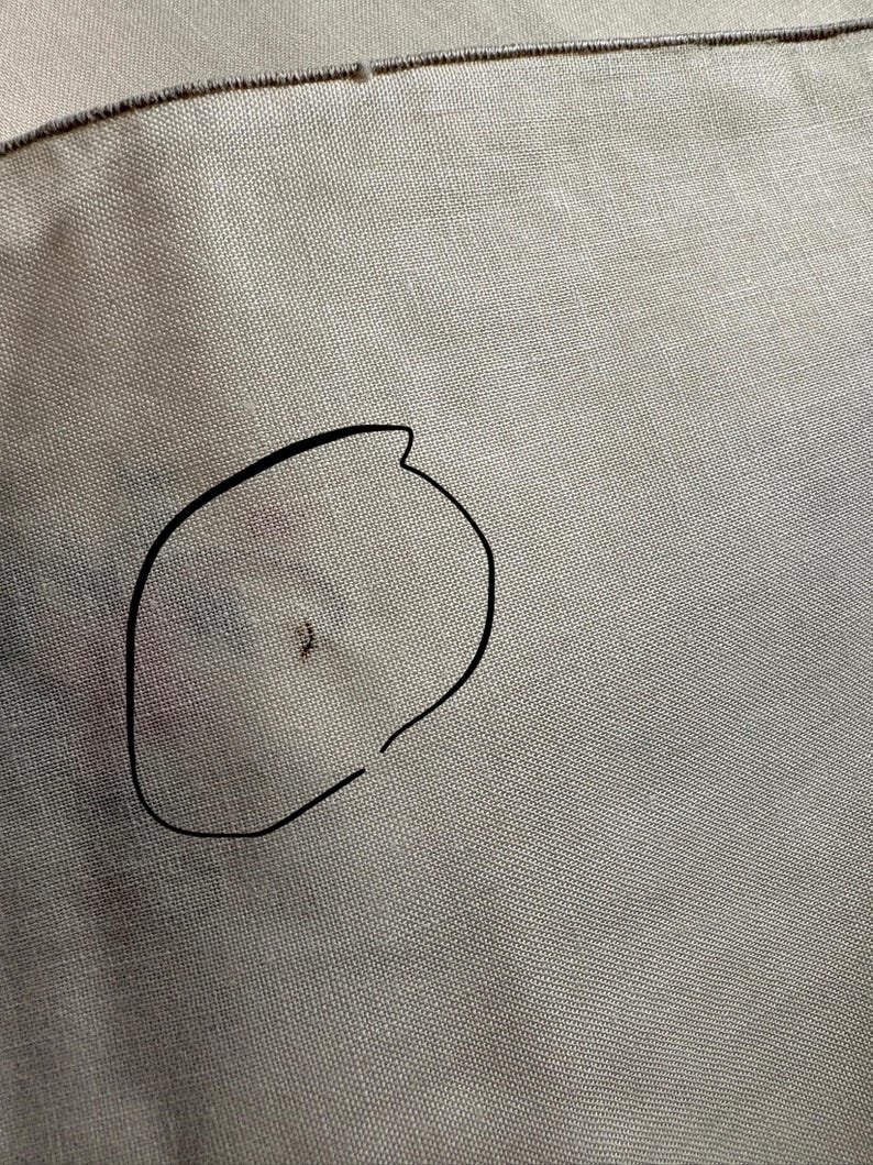 May include: Close-up of a beige fabric with a black outline of a shape drawn on it. The fabric has a woven texture and a small dark spot within the outline. A brown thread is visible at the top.