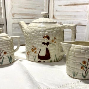 May include: A vintage tea set comprising a white teapot, creamer, and sugar bowl. Each piece has a stone-like texture and a floral design with a girl in a brown dress.