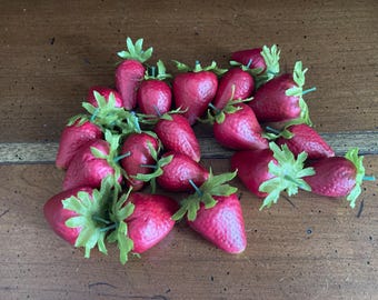 Puede incluir: Un grupo de 15 fresas rojas artificiales con hojas verdes. Las fresas están dispuestas en una pila sobre una superficie de madera.