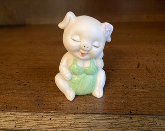Puede incluir: Una figurita de cerdo de cerámica blanca que lleva un traje de baño verde con lunares blancos y una flor rosa en la oreja. El cerdo sonríe con los ojos cerrados.