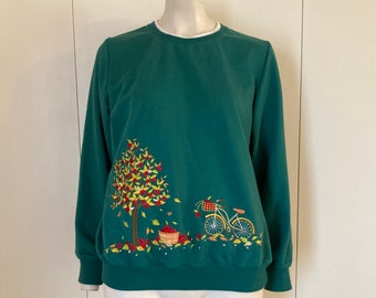 Puede incluir: Una sudadera de color verde azulado con un diseño bordado de un árbol con hojas rojas y amarillas, una cesta de manzanas y una bicicleta con una cesta.