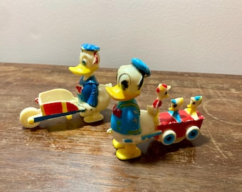 Puede incluir: Dos figuras vintage de plástico de Donald Duck. Un Donald está empujando una carretilla de juguete roja y azul. El otro Donald está tirando de un vagón de juguete rojo y azul con dos figuras más pequeñas de Donald Duck dentro.