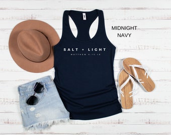 May include: A navy blue racerback tank top with the text "SALT + LIGHT" and "MATTHEW 5:13-16" in white. The tank top is paired with denim shorts, a brown hat, sunglasses, and wooden sandals.