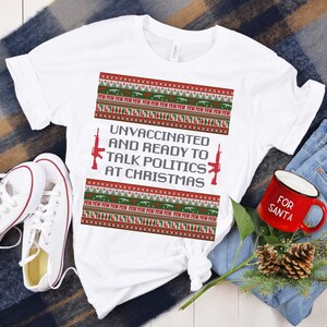 May include: White t-shirt with a festive design featuring red and green patterns, guns, and the text "UNVACCINATED AND READY TO TALK POLITICS AT CHRISTMAS." The shirt is paired with white sneakers, blue jeans, and a red mug.