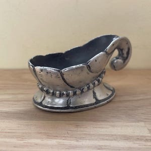May include: A small, antique silver-colored dish with a decorative, curved handle. The dish has a scalloped rim and a beaded base. The interior of the dish is dark. The dish is likely made of metal.