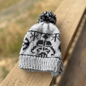 May include: A gray knit beanie with a black and white pom-pom on top. The hat features a black Darth Vader design and a ribbed brim. The hat is made of a soft, knitted material.