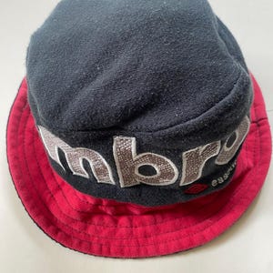 May include: A black bucket hat with a red brim. The hat has embroidered lettering in a light brown colour. The brand name is visible on the hat. The hat is made of a soft fabric.