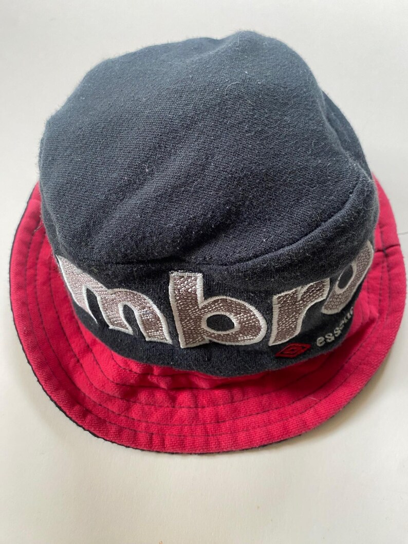 May include: A black bucket hat with a red brim. The hat has embroidered lettering in a light brown colour. The brand name is visible on the hat. The hat is made of a soft fabric.