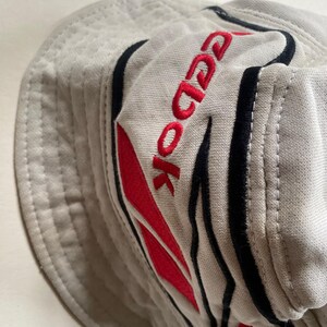 May include: A light grey bucket hat with the brand name "Reebok" embroidered in red. The hat features black and red accents and a stitched brim. The hat is made of a soft, textured fabric.