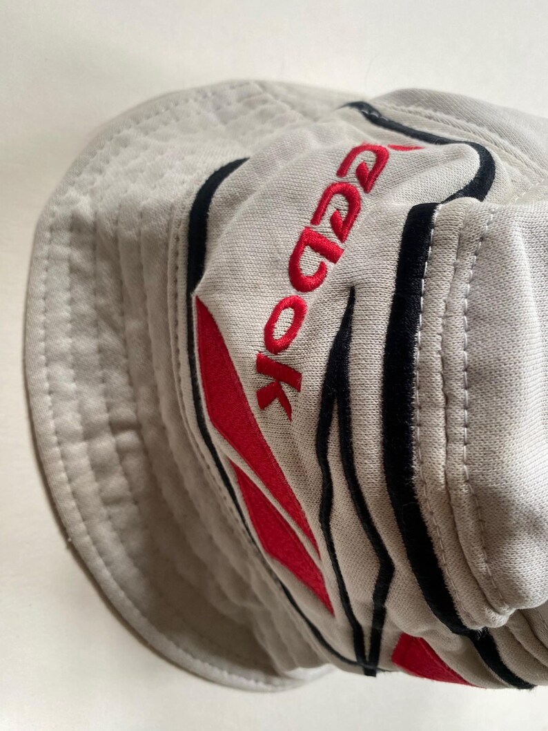 May include: A light grey bucket hat with the brand name "Reebok" embroidered in red. The hat features black and red accents and a stitched brim. The hat is made of a soft, textured fabric.