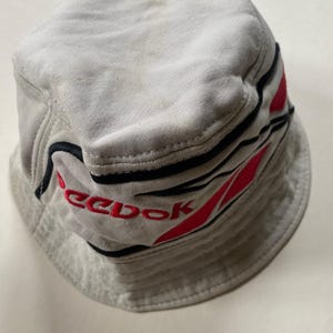 May include: A light grey bucket hat with the brand name "Reebok" embroidered in red. The hat has black and red stripes around the brim. The hat is made of a soft material, possibly cotton.