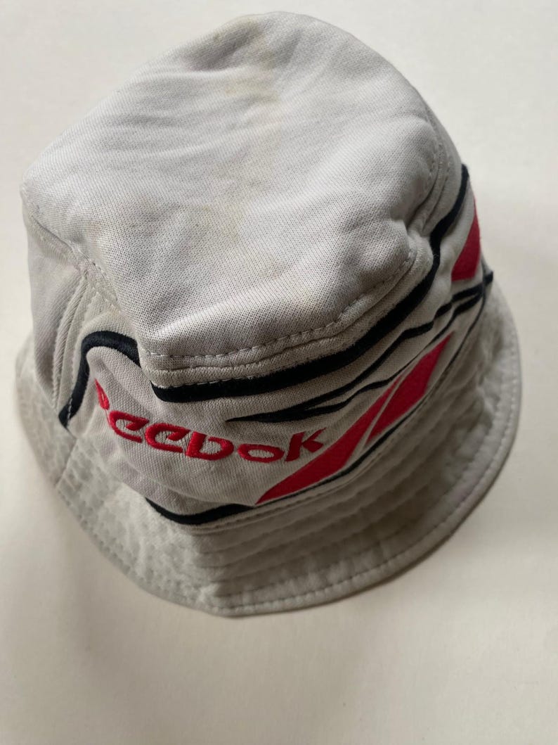 May include: A light grey bucket hat with the brand name "Reebok" embroidered in red. The hat has black and red stripes around the brim. The hat is made of a soft material, possibly cotton.