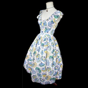 May include: A vintage-style dress on a mannequin. The dress features a white background with a repeating pattern of blue, green, and yellow figures. It has a ruffled neckline and cap sleeves. The dress is knee-length and has a full skirt.