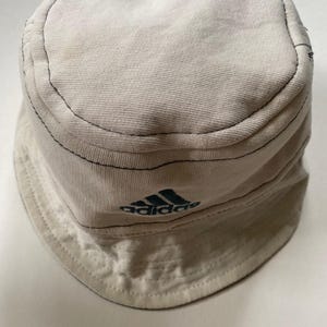 May include: Off-white bucket hat featuring the Adidas logo in teal. The hat has a rounded crown and a downward-sloping brim. Black stitching outlines the seams. The hat is made of a soft, textured fabric.