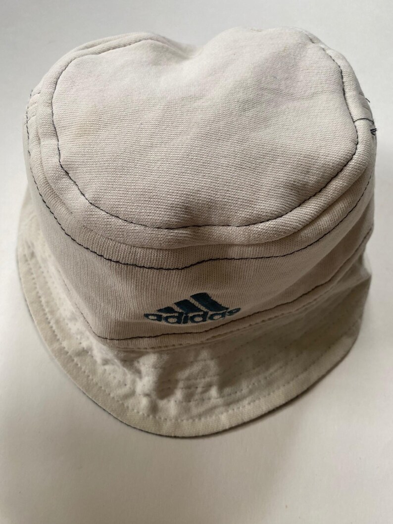 May include: Off-white bucket hat featuring the Adidas logo in teal. The hat has a rounded crown and a downward-sloping brim. Black stitching outlines the seams. The hat is made of a soft, textured fabric.