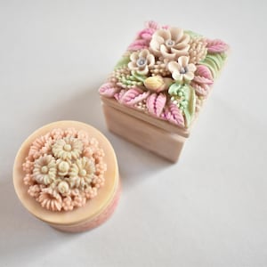 Puede incluir: Dos cajas decorativas con diseños florales. Una es rectangular con tapa, y la otra es ovalada. Ambas son de color beige claro con detalles florales en rosa pastel, verde y blanco. Las cajas están sobre una superficie blanca.