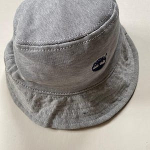 May include: A grey bucket hat with a small, dark blue embroidered logo. The hat is made of a soft, textured fabric with white stitching along the seams. The brim is slightly curved. A casual hat for everyday wear.