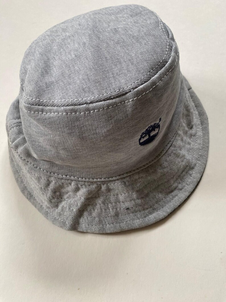 May include: A grey bucket hat with a small, dark blue embroidered logo. The hat is made of a soft, textured fabric with white stitching along the seams. The brim is slightly curved. A casual hat for everyday wear.