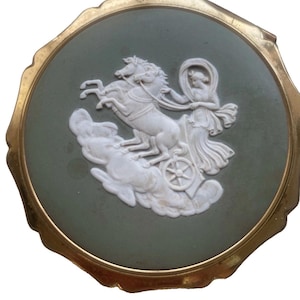May include: A round compact mirror featuring a sage green center inlay with a white relief carving of a figure driving a horse-drawn chariot through clouds. The circular piece is framed by a scalloped gold-toned metal border with a smooth finish.