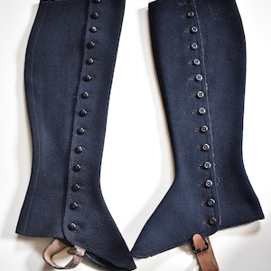 May include: A pair of dark navy blue wool gaiters featuring a vertical row of small black buttons along the outer edge. Each gaiter includes a tan leather strap with a metal buckle at the bottom to secure the garment around the lower leg area.