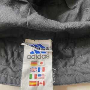 May include: Close-up of a black Adidas bucket hat. The hat's fabric is textured. A white tag with the Adidas logo and flags of various countries is visible.