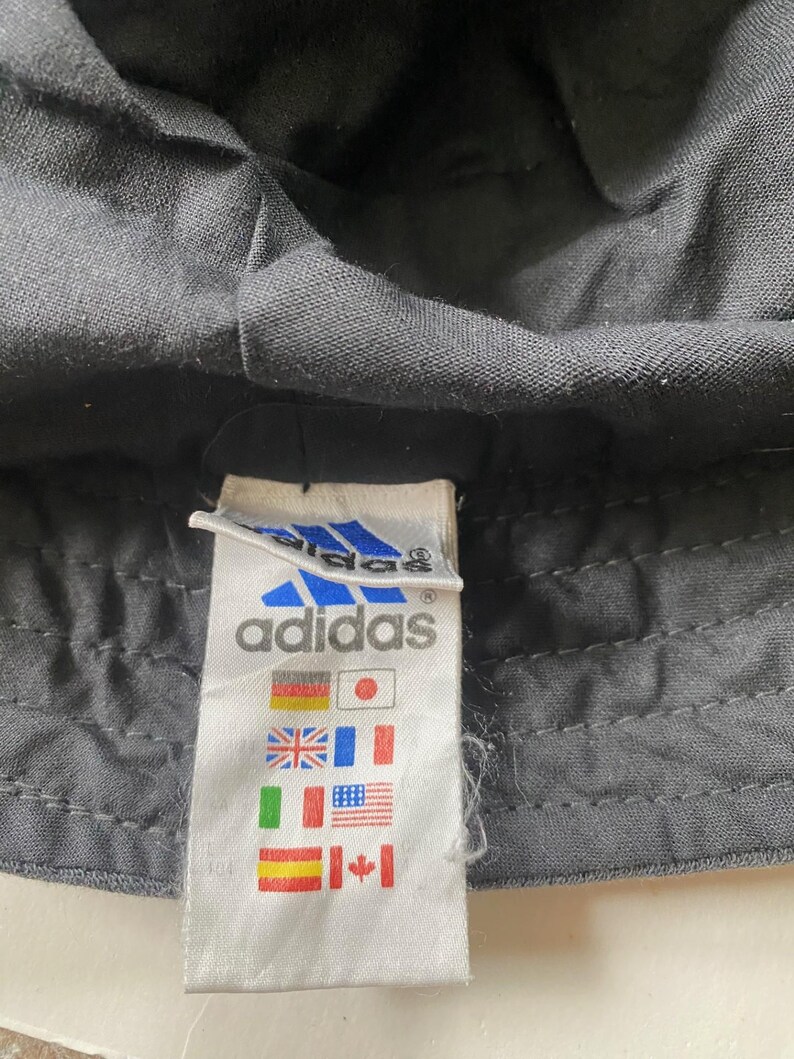May include: Close-up of a black Adidas bucket hat. The hat's fabric is textured. A white tag with the Adidas logo and flags of various countries is visible.