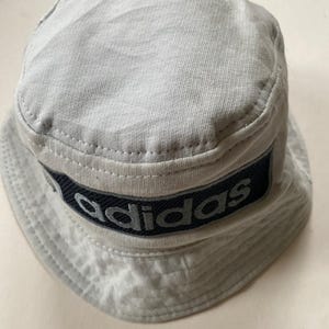 May include: A light grey bucket hat with a dark blue band displaying the brand name "adidas" in white lettering. The hat is made of a soft fabric and has a classic, casual style. The hat is a light grey colour.