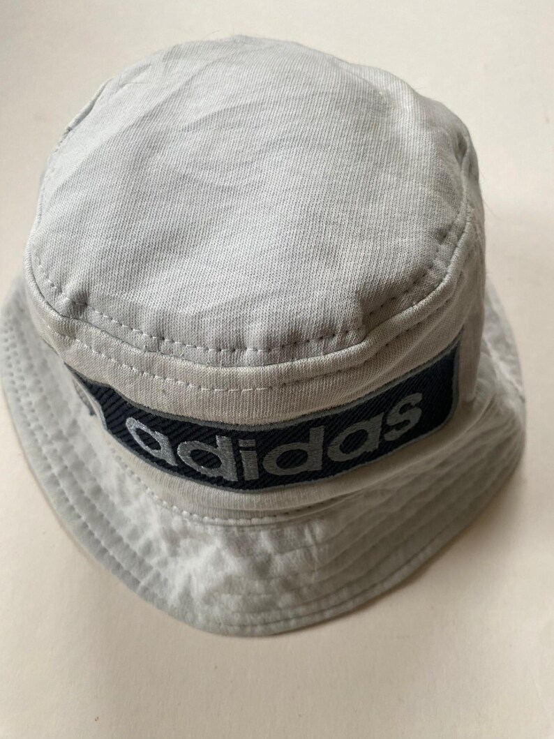 May include: A light grey bucket hat with a dark blue band displaying the brand name "adidas" in white lettering. The hat is made of a soft fabric and has a classic, casual style. The hat is a light grey colour.