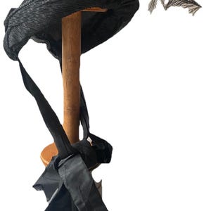 May include: A black pleated fabric hat with a decorative feather detail. The hat is displayed on a wooden stand, with long fabric ties hanging down. The hat has a vintage or retro style.
