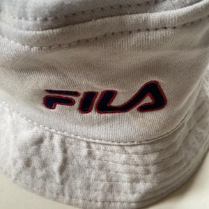 May include: Close-up of a light grey bucket hat with the FILA logo embroidered in navy blue and red. The hat is made of a soft, textured fabric and features a classic bucket hat design.