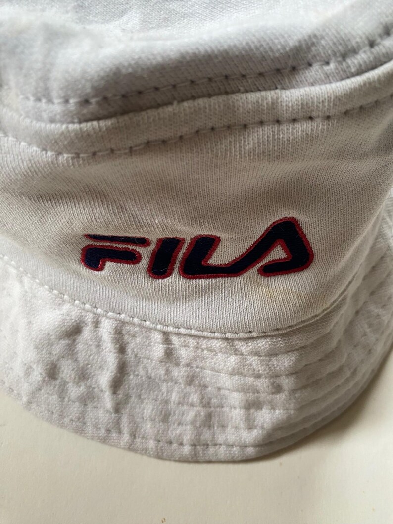 May include: Close-up of a light grey bucket hat with the FILA logo embroidered in navy blue and red. The hat is made of a soft, textured fabric and features a classic bucket hat design.