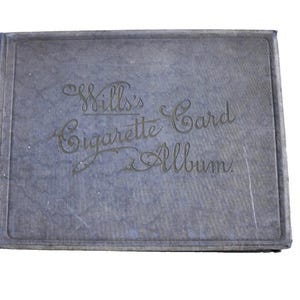 May include: A rectangular, dark blue, textured album with a fabric-like cover. The front cover features the embossed text "Wills's Cigarette Card Album" in a decorative, cursive script centered on the front of the book.