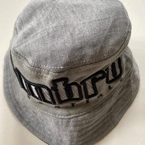 May include: A grey bucket hat with the word "Unbro" embroidered in black across the front. The hat is made of a soft fabric and features a rounded crown and a downward-sloping brim. The hat is a casual accessory.