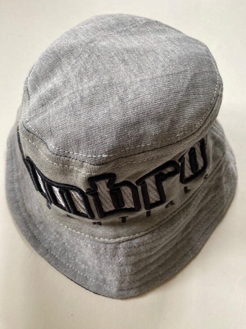 May include: A grey bucket hat with the word "Unbro" embroidered in black across the front. The hat is made of a soft fabric and features a rounded crown and a downward-sloping brim. The hat is a casual accessory.