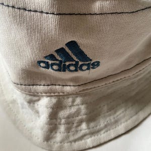 May include: A light beige bucket hat featuring the Adidas logo embroidered in dark blue. The hat has a stitched brim and dark blue stitching along the seams. The hat is made of a soft, textured fabric.