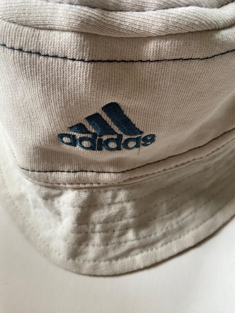 May include: A light beige bucket hat featuring the Adidas logo embroidered in dark blue. The hat has a stitched brim and dark blue stitching along the seams. The hat is made of a soft, textured fabric.