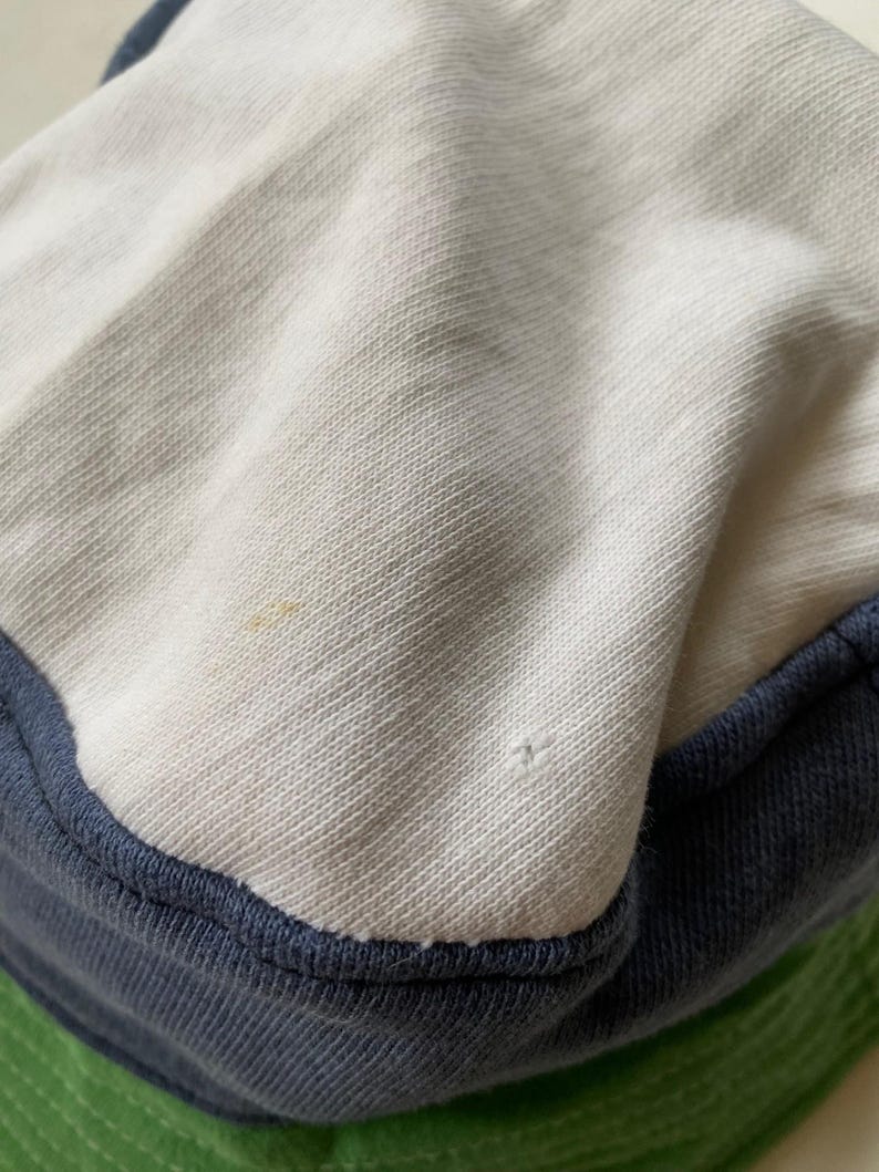 May include: Close-up of a white and blue bucket hat. The hat has a white crown, a navy blue brim, and a green underside. The fabric appears to be a soft, textured material.