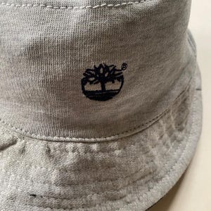 May include: A light grey bucket hat with a black embroidered tree logo. The hat is made of a soft, textured fabric and features a stitched brim. The Timberland logo is on the front.