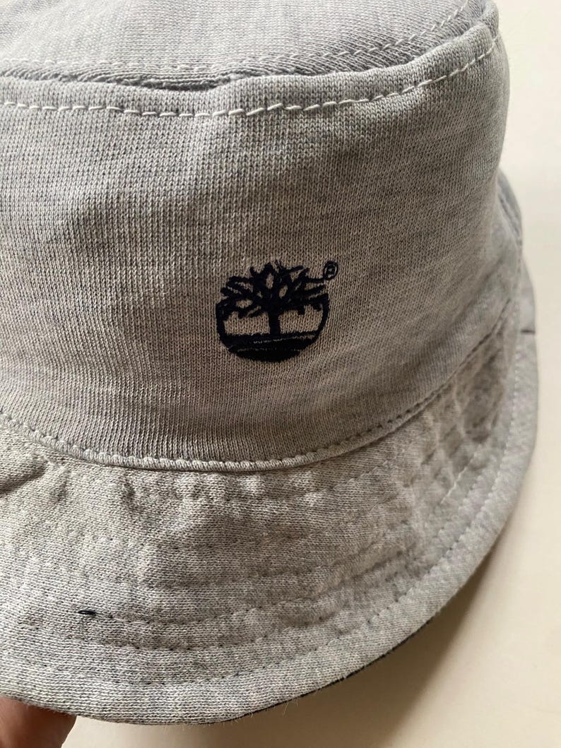 May include: A light grey bucket hat with a black embroidered tree logo. The hat is made of a soft, textured fabric and features a stitched brim. The Timberland logo is on the front.