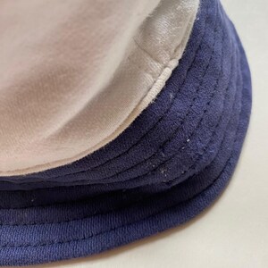 May include: A close-up of a three-tone bucket hat. The hat features a white top, a light grey middle section, and a navy blue brim. The hat is made of a soft, textured fabric.