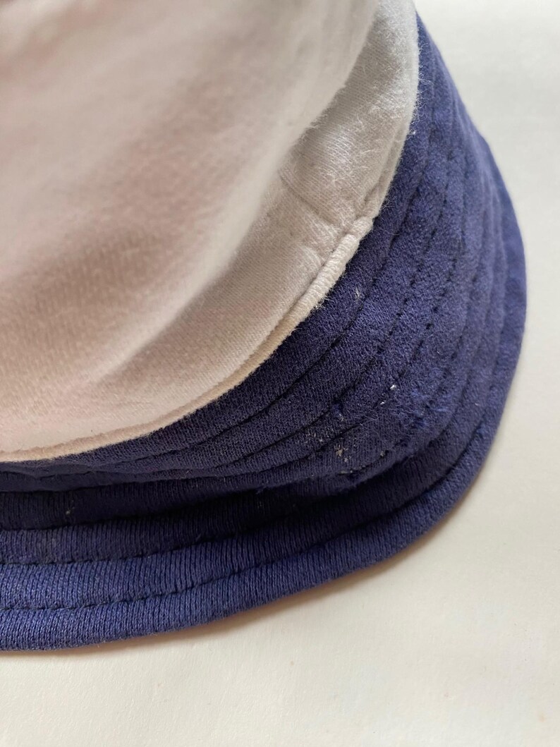 May include: A close-up of a three-tone bucket hat. The hat features a white top, a light grey middle section, and a navy blue brim. The hat is made of a soft, textured fabric.