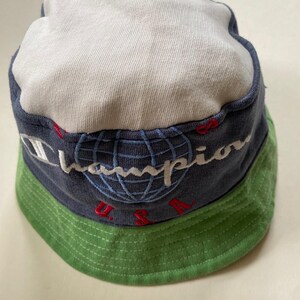 May include: A Champion bucket hat featuring a white crown, a navy blue band with the Champion logo and globe design, and a green brim. The text "U.S.A." is embroidered in red. A casual, sporty accessory.