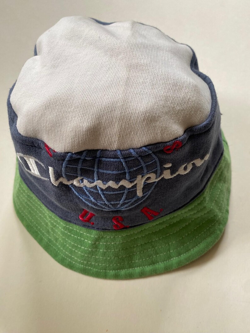 May include: A Champion bucket hat featuring a white crown, a navy blue band with the Champion logo and globe design, and a green brim. The text "U.S.A." is embroidered in red. A casual, sporty accessory.