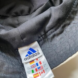 May include: Close-up of an Adidas bucket hat. The hat is black on the inside and off-white on the outside. A white tag with the Adidas logo and international flags is visible.