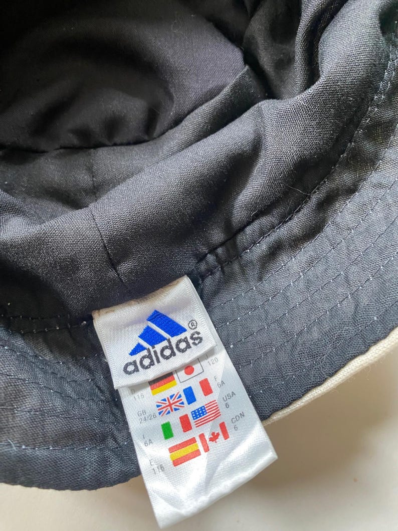 May include: Close-up of an Adidas bucket hat. The hat is black on the inside and off-white on the outside. A white tag with the Adidas logo and international flags is visible.