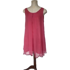 May include: A sleeveless, knee-length pink dress with a scoop neckline and three buttons. The dress has a flowing, layered skirt with a scalloped hem. It is displayed on a mannequin with a wooden stand.