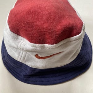 May include: A multi-coloured bucket hat with a red top, white mid-section, and navy blue brim. The hat features a small orange Nike swoosh logo on the white section. This casual hat is made of a soft fabric.