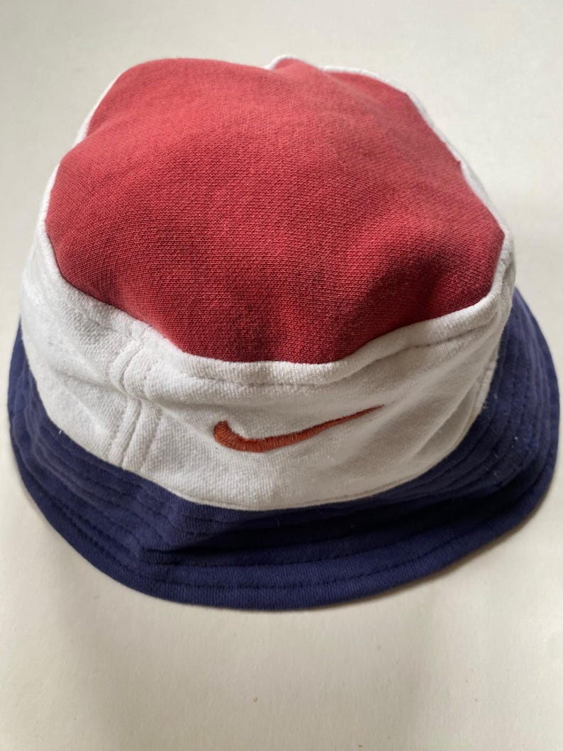 May include: A multi-coloured bucket hat with a red top, white mid-section, and navy blue brim. The hat features a small orange Nike swoosh logo on the white section. This casual hat is made of a soft fabric.