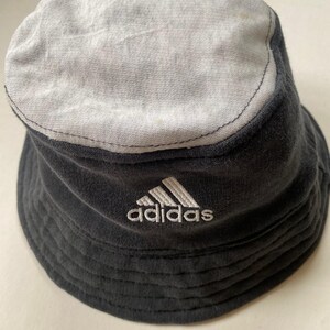 May include: A black and grey bucket hat with the Adidas logo embroidered in white. The hat has a two-tone design, with a grey top and a black body. The hat is made of a soft, textured fabric.