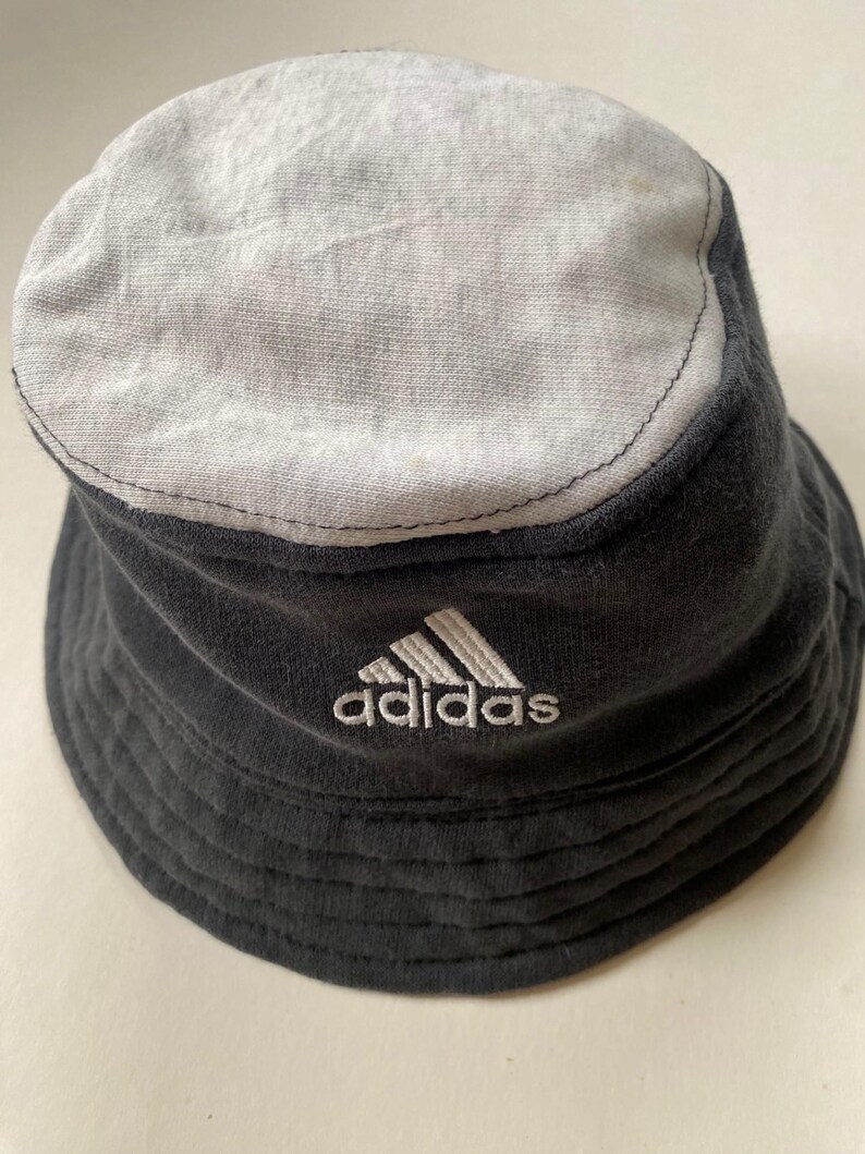 May include: A black and grey bucket hat with the Adidas logo embroidered in white. The hat has a two-tone design, with a grey top and a black body. The hat is made of a soft, textured fabric.